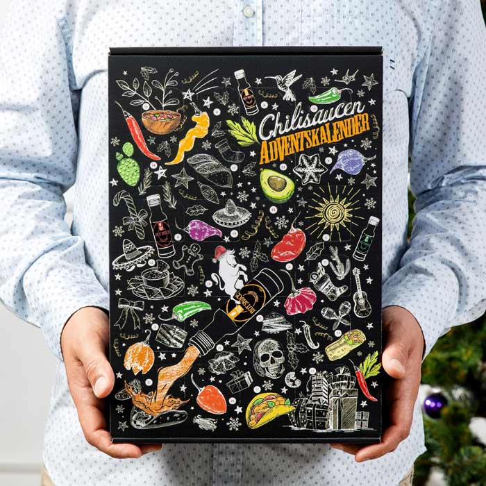 Eine Person in einem hellblauen Hemd hält einen Chilisaucen-Adventskalender. Die Verpackung ist schwarz und mit bunten Illustrationen verziert, darunter Chilischoten, Avocados, Totenköpfe und verschiedene Lebensmittel.