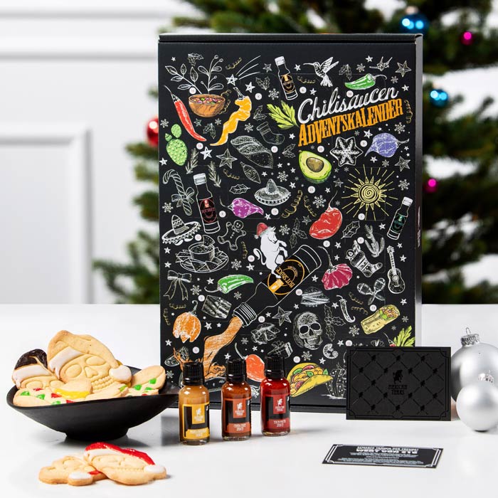 Ein Chilisaucen-Adventskalender steht vor einem weihnachtlich dekorierten Hintergrund. Die Verpackung ist schwarz und bunt illustriert mit Motiven wie Chilischoten, Avocado, Totenköpfen und anderen Lebensmitteln. Vor dem Kalender stehen drei kleine Flaschen mit verschiedenen Chilisaucen. Neben den Flaschen liegt ein Teller mit Weihnachtsplätzchen und verschiedene Dekorationsartikel, darunter Christbaumkugeln und eine Geschenkkarte. Im Hintergrund ist ein geschmückter Weihnachtsbaum zu sehen.