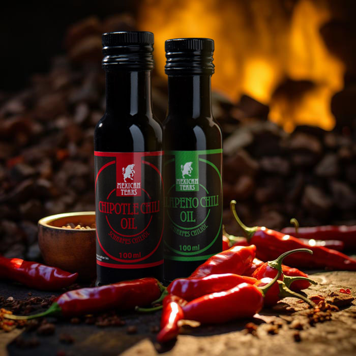 Zwei Flaschen von Mexican Tears Chiliöl, eine mit Chipotle Chili Öl und die andere mit Jalapeño Chili Öl, stehen im Vordergrund auf einem rustikalen Untergrund mit roten Chilischoten drumherum. Im Hintergrund flackert ein warmes Feuer, was die gemütliche Atmosphäre und die Wärme der Produkte unterstreicht.