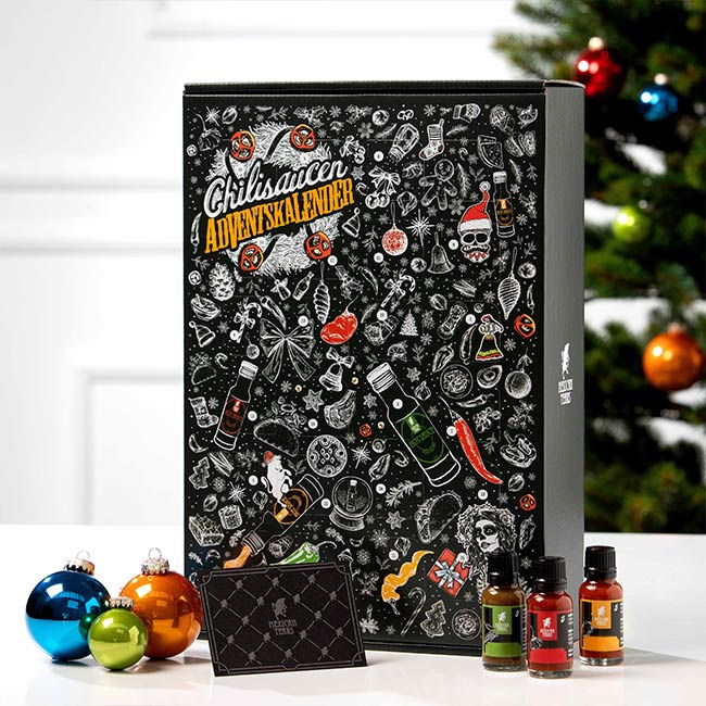 Ein Chilisaucen-Adventskalender steht vor einem weihnachtlich dekorierten Hintergrund. Die Verpackung ist schwarz und bunt illustriert mit Motiven wie Chilischoten, Avocado, Totenköpfen und anderen Lebensmitteln. Vor dem Kalender stehen drei kleine Flaschen mit verschiedenen Chilisaucen. Neben den Flaschen liegt ein Teller mit Weihnachtsplätzchen und verschiedene Dekorationsartikel, darunter Christbaumkugeln und eine Geschenkkarte. Im Hintergrund ist ein geschmückter Weihnachtsbaum zu sehen.