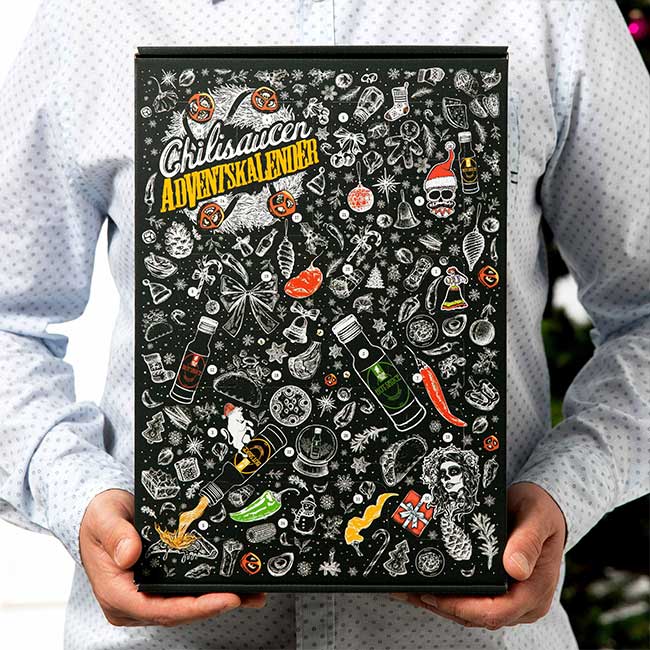 Eine Person in einem hellblauen Hemd hält einen Chilisaucen-Adventskalender. Die Verpackung ist schwarz und mit bunten Illustrationen verziert, darunter Chilischoten, Avocados, Totenköpfe und verschiedene Lebensmittel.