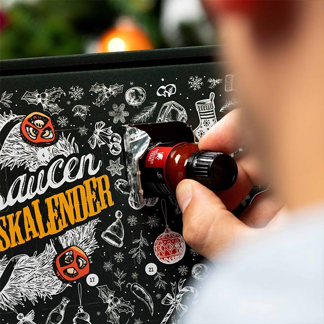 Hand nimmt Chilisauce aus Chilisaucen-Adventskalenders und zeigt eine Übersicht der verschiedenen Chili-Sorten und ihrer Schärfegrade. Oben ist das Logo von Mexican Tears.