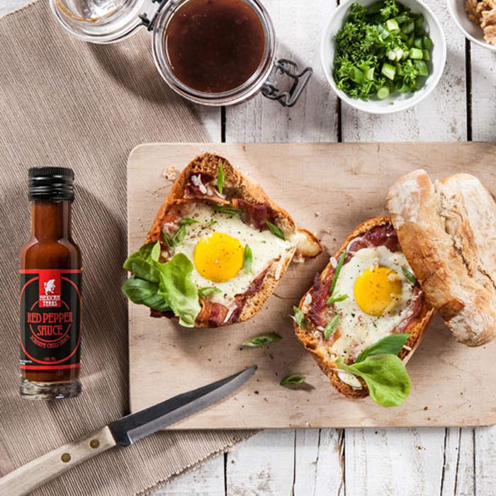 Red Pepper Sauce Frühstück - Ein pikantes Start in den Tag. Zwei herzhafte belegte Brote mit Spiegelei, grünem Salat und knusprigem Speck auf einem Holzbrett, begleitet von einer Flasche Red Pepper Sauce und frischen Kräutern im Hintergrund.