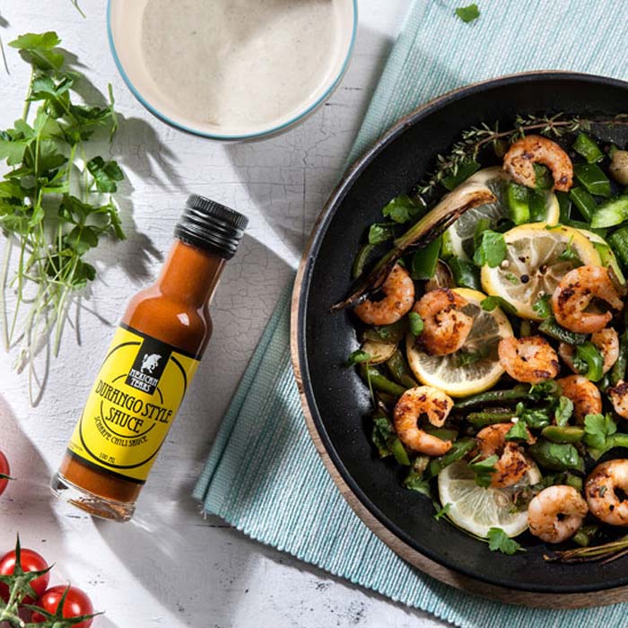 Durango Style Chili Sauce: Der perfekte Partner für Meeresfrüchte und Gemüse. Eine Pfanne mit saftigen, gebratenen Garnelen und knackigem grünen Gemüse, garniert mit Zitronenscheiben und frischen Kräutern, neben einer Flasche Durango Style Chili Sauce auf einer hellen Arbeitsfläche mit blauem Geschirrtuch.
