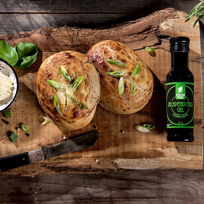Gebackenes Brot und Jalapeno Chili Öl - Perfekte Kombination für ein würziges Geschmackserlebnis Rustikales Arrangement mit zwei frisch gebackenen Broten und einer Flasche Jalapeno Chili Öl auf einem Holzbrett, begleitet von Käsescheiben und frischem Basilikum.