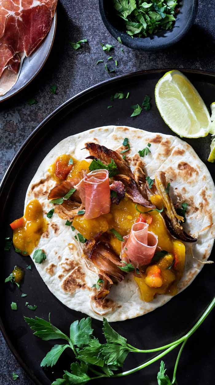 Herzhaftes Taco-Gericht - Perfekt für Abendbrot Taco auf dunklem Broste Teller mit Mango Chutney, Serrano Schinken und Pulled Pork. Dazu Beilagen wie Koriander und Limetten. Von oben fotografiert.