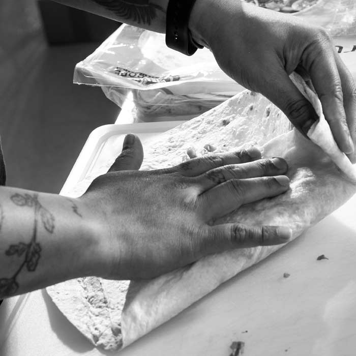 Eine Person faltet eine Weizentortilla mit Pulled Chicken und Käsefüllung zu einem Crunchwrap, erkennbar an den tätowierten Unterarmen und geschickten Händen.